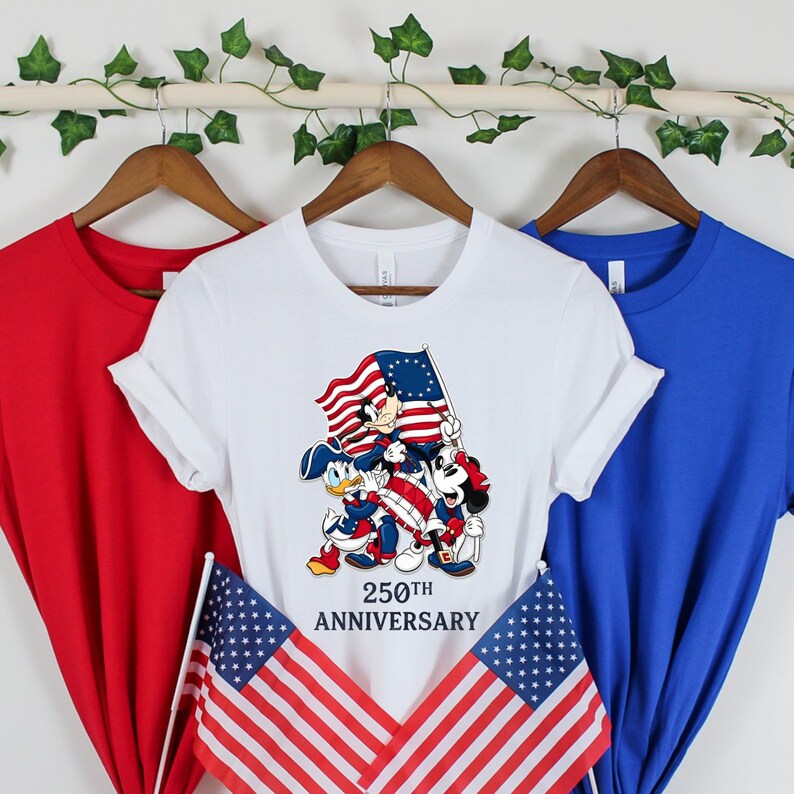May include: Three t-shirts in red, white, and blue hang on a wooden rod. The white t-shirt features a graphic of Mickey Mouse, Donald Duck, and Goofy holding a flag with the text "250th Anniversary." Two American flags are in the foreground.
