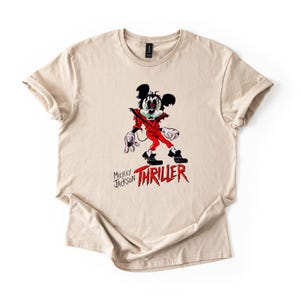 May include: A beige short-sleeve t-shirt featuring a graphic of a cartoon mouse dressed in a red jacket, black trousers, and white gloves. The graphic includes the text "MICKEY JACKSON" and the word "THRILLER" in a bold, red, jagged font.