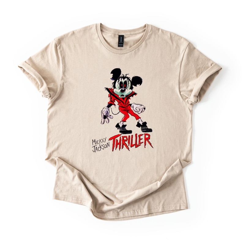May include: A beige short-sleeve t-shirt featuring a graphic of a cartoon mouse dressed in a red jacket, black trousers, and white gloves. The graphic includes the text "MICKEY JACKSON" and the word "THRILLER" in a bold, red, jagged font.