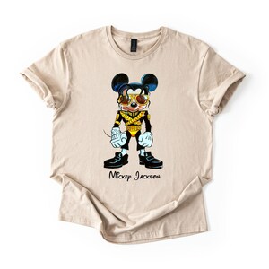 Puede incluir: Una camiseta beige de manga corta con un gr&aacute;fico de un rat&oacute;n de dibujos animados vestido con una chaqueta militar negra y dorada con gafas de sol doradas. El personaje tiene el pelo oscuro, guantes blancos y zapatos negros. El texto Mickey Jackson est&aacute; impreso debajo del gr&aacute;fico en una fuente cursiva negra.
