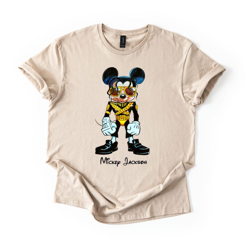 Puede incluir: Una camiseta beige de manga corta con un gr&aacute;fico de un rat&oacute;n de dibujos animados vestido con una chaqueta militar negra y dorada con gafas de sol doradas. El personaje tiene el pelo oscuro, guantes blancos y zapatos negros. El texto Mickey Jackson est&aacute; impreso debajo del gr&aacute;fico en una fuente cursiva negra.