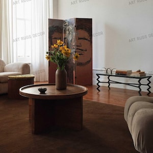 May include: A living room scene featuring a round wooden coffee table, a decorative screen with a face design, and a vase of yellow flowers. The room has a brown rug and a modern black metal and glass table. The text "ART RUGS DECOR" is visible.