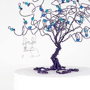 May include: A purple wire tree with blue crystal accents and a silver wire couple on a swing. The tree is on a white cake.