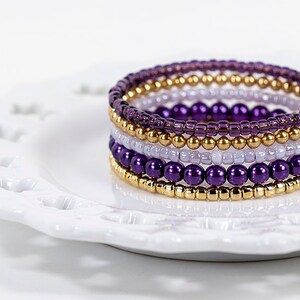 Könnte beinhalten: Ein mehrlagiges Armband mit lila und goldenen Perlen. Das Armband besteht aus mehreren Perlenreihen, wobei die lila Perlen am auffälligsten sind. Das Armband liegt auf einem weißen Teller.