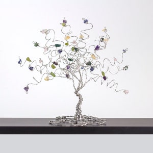 May include: A silver wire tree sculpture with colorful gemstone leaves. The tree is on a dark brown surface.