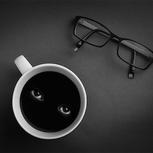 May include: A white coffee mug with a black liquid inside. Two eyes are visible in the liquid. A pair of black framed glasses are lying on a dark surface.