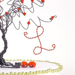 Puede incluir: Una escultura de &aacute;rbol de alambre con cuentas rojas y una letra L de alambre rojo colgando de las ramas. Dos cuentas de color naranja en forma de calabaza con caras sonrientes est&aacute;n sobre una base blanca con un borde de cuentas doradas.