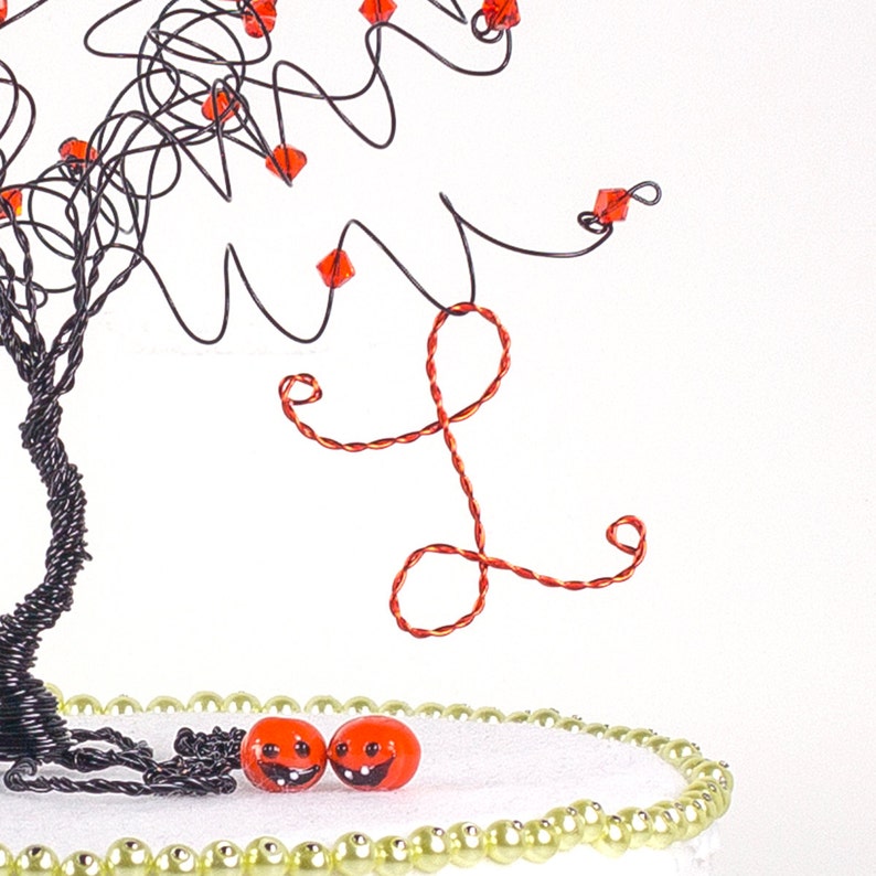 Puede incluir: Una escultura de &aacute;rbol de alambre con cuentas rojas y una letra L de alambre rojo colgando de las ramas. Dos cuentas de color naranja en forma de calabaza con caras sonrientes est&aacute;n sobre una base blanca con un borde de cuentas doradas.