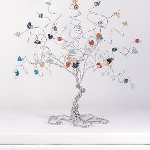 May include: A silver wire tree sculpture with colorful gemstone leaves. The tree is on a white surface.