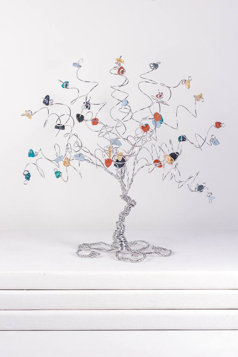 May include: A silver wire tree sculpture with colorful gemstone leaves. The tree is on a white surface.