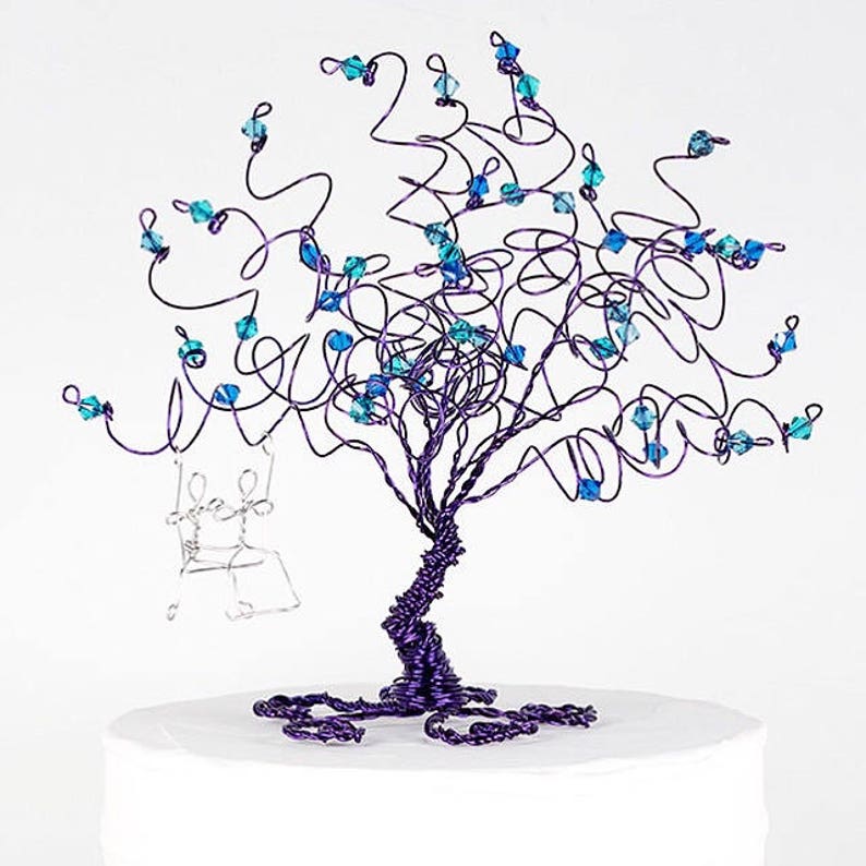 May include: A purple wire tree with blue crystal accents and a silver wire couple on a swing. The tree is on a white cake.