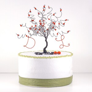 Puede incluir: Un &aacute;rbol de alambre negro con cuentas rojas y las letras D y L en las ramas. El &aacute;rbol est&aacute; sobre un pastel blanco con una cinta verde y cuentas doradas alrededor de la base.