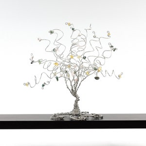May include: A silver wire tree sculpture with a twisted trunk and branches adorned with colorful gemstones. The tree is displayed on a black shelf.