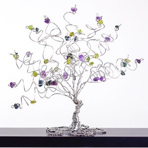 May include: A silver wire tree sculpture with purple, green, and blue gemstones attached to the branches. The tree is on a black surface.