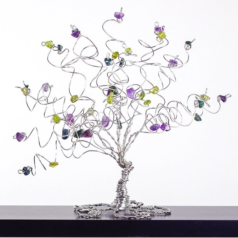 May include: A silver wire tree sculpture with purple, green, and blue gemstones attached to the branches. The tree is on a black surface.
