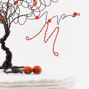 Puede incluir: Una escultura de alambre negro de un &aacute;rbol con cuentas rojas y adornos de calabaza naranja. El &aacute;rbol est&aacute; sobre una superficie blanca.