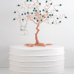 May include: A copper wire tree with teal beads and a silver wire swing hanging from the branches. The tree is on top of a white cake.