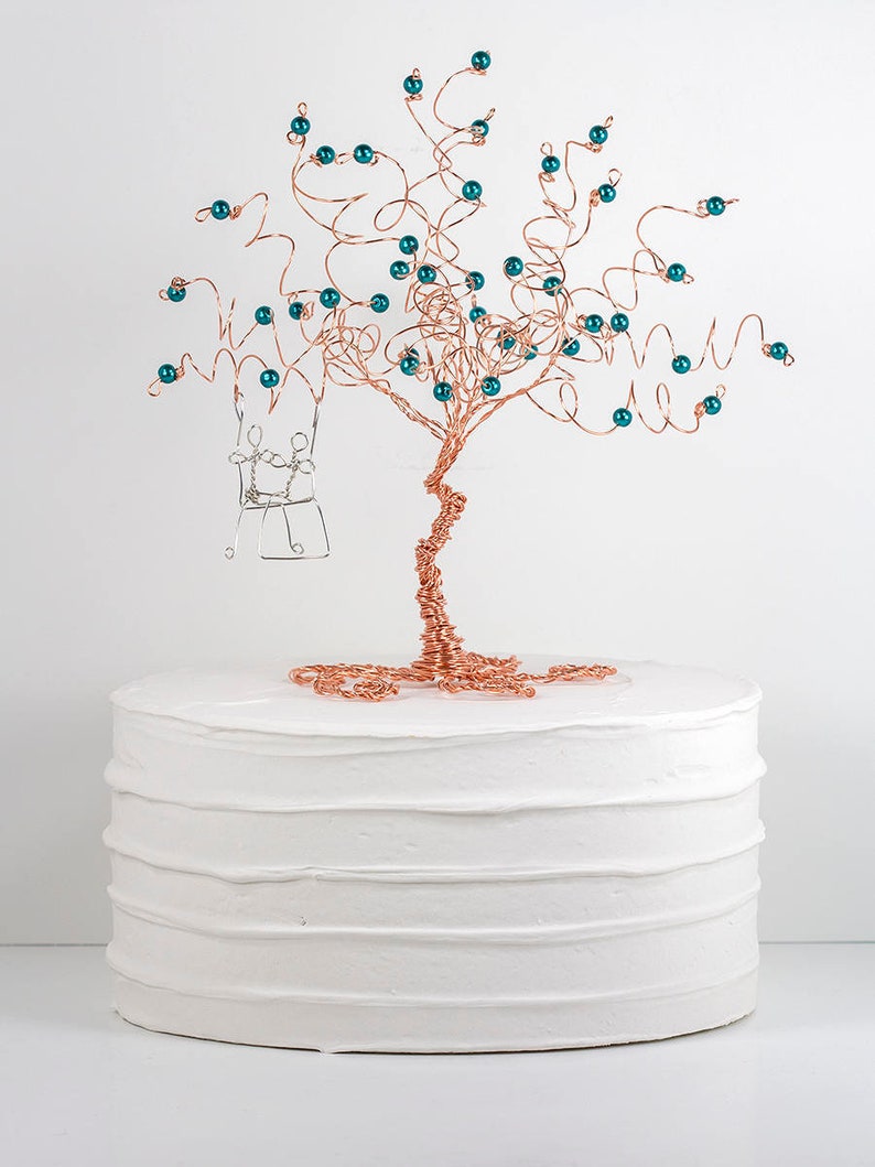 May include: A copper wire tree with teal beads and a silver wire swing hanging from the branches. The tree is on top of a white cake.