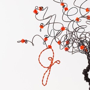 Puede incluir: Una escultura de alambre de un &aacute;rbol con cuentas rojas para las hojas. El &aacute;rbol est&aacute; hecho de alambre negro y las cuentas son de un color rojo anaranjado brillante. El &aacute;rbol est&aacute; de pie sobre una superficie blanca.