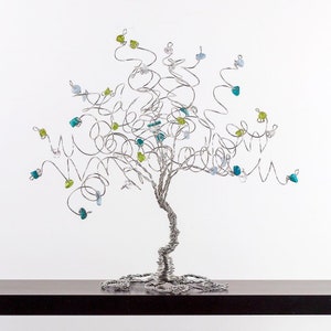 May include: A silver wire tree sculpture with blue, green, and clear glass beads. The tree is on a dark brown surface.