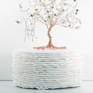 May include: A white cake with a wire tree topper decorated with green beads and a small wire swing. The cake is made of multiple layers of white frosting.