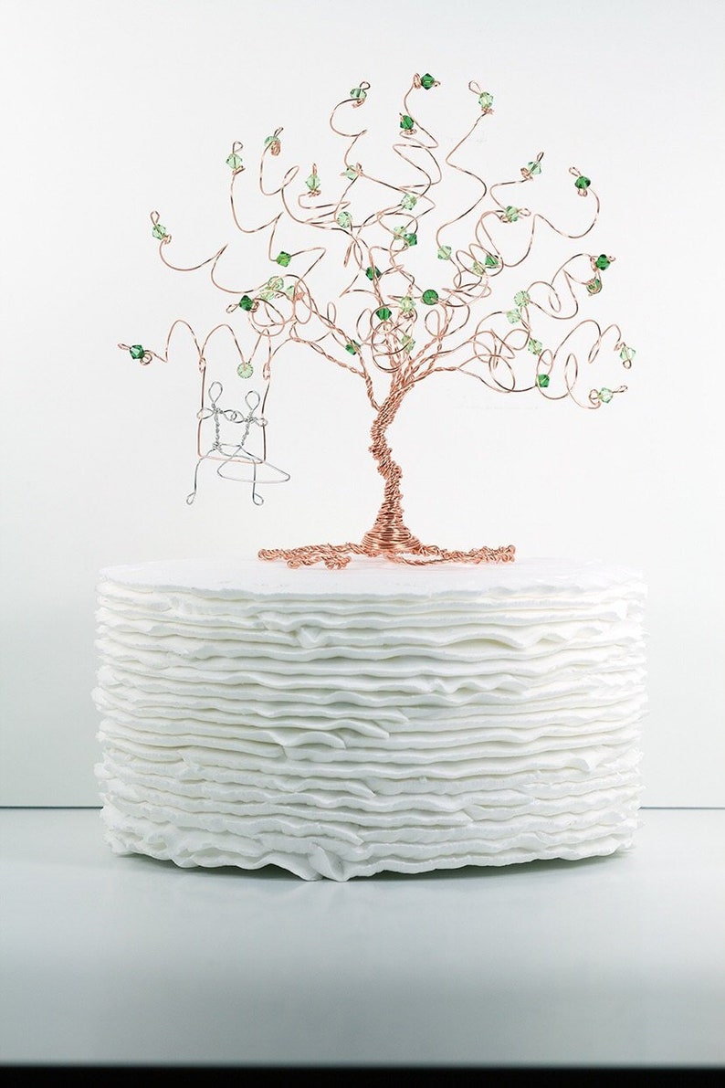 May include: A white cake with a wire tree topper decorated with green beads and a small wire swing. The cake is made of multiple layers of white frosting.
