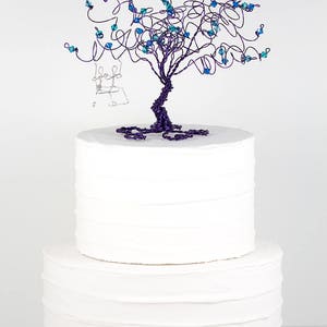 May include: A white two-tiered wedding cake topped with a purple wire tree sculpture with blue crystal beads. The tree has a small wire couple sitting on a swing.