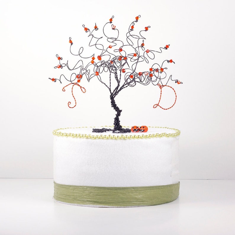 Puede incluir: Un &aacute;rbol de alambre negro con cuentas naranjas para las hojas, sentado sobre un pastel blanco con una cinta verde y un borde de cuentas doradas. Dos peque&ntilde;as calabazas naranjas est&aacute;n en la base del &aacute;rbol.