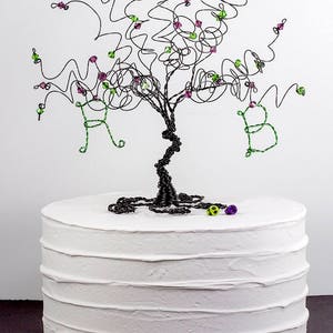 Puede incluir: Un adorno para pastel de alambre negro con forma de &aacute;rbol con cuentas verdes y moradas. El &aacute;rbol tiene las letras "R" y "B" formadas en las ramas. El adorno est&aacute; sobre un pastel blanco.
