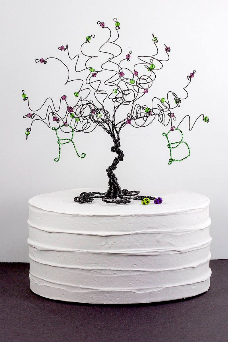 Puede incluir: Un adorno para pastel de alambre negro con forma de &aacute;rbol con cuentas verdes y moradas. El &aacute;rbol tiene las letras "R" y "B" formadas en las ramas. El adorno est&aacute; sobre un pastel blanco.