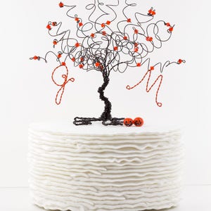 Puede incluir: Un pastel blanco con un adorno de &aacute;rbol de alambre negro decorado con cuentas rojas. El &aacute;rbol tiene dos adornos en forma de calabaza roja en la base.