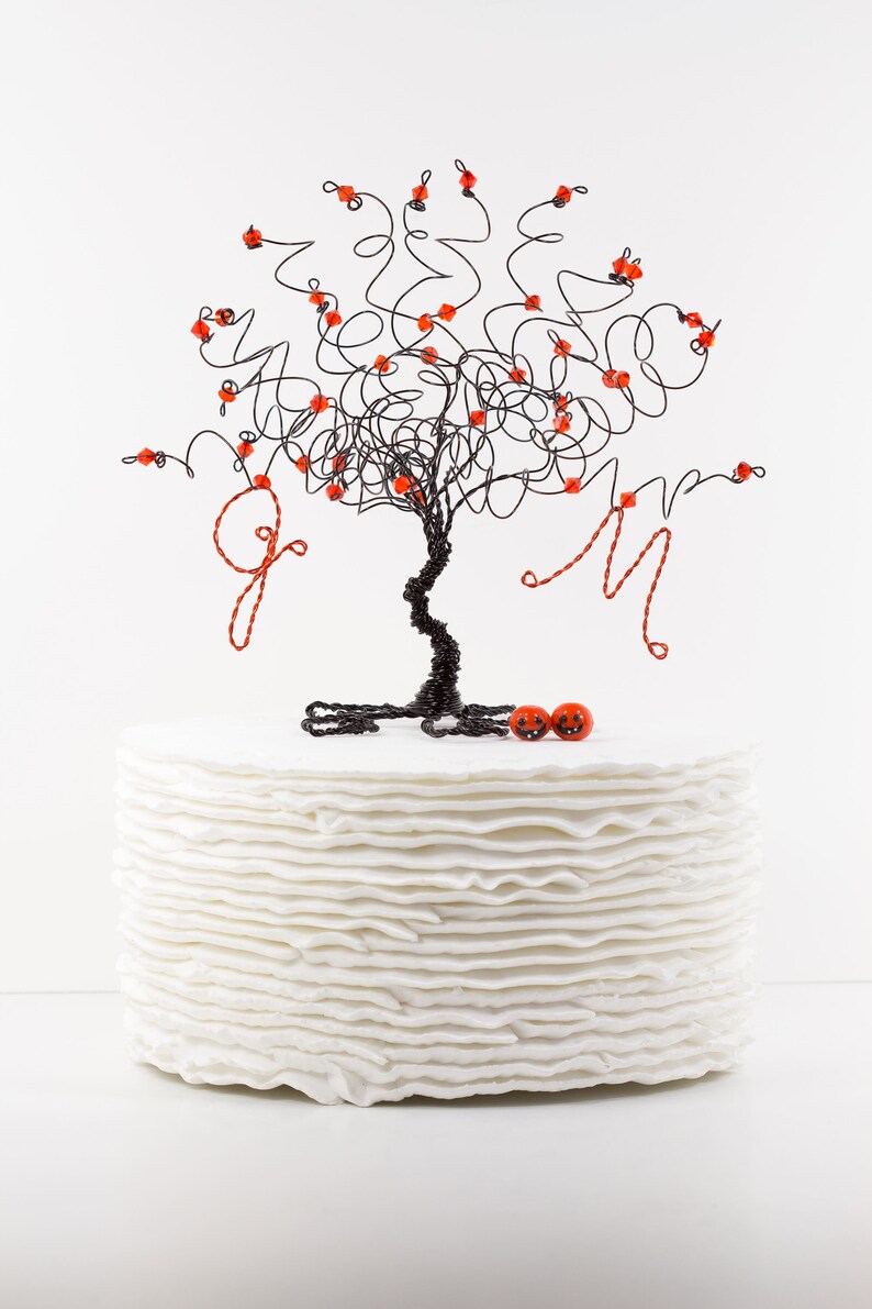 Puede incluir: Un pastel blanco con un adorno de &aacute;rbol de alambre negro decorado con cuentas rojas. El &aacute;rbol tiene dos adornos en forma de calabaza roja en la base.