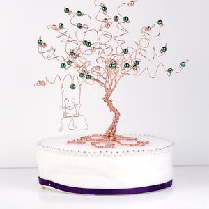 May include: A copper wire tree sculpture with green beads and a wire couple on a swing. The tree is on a white base with a purple ribbon.