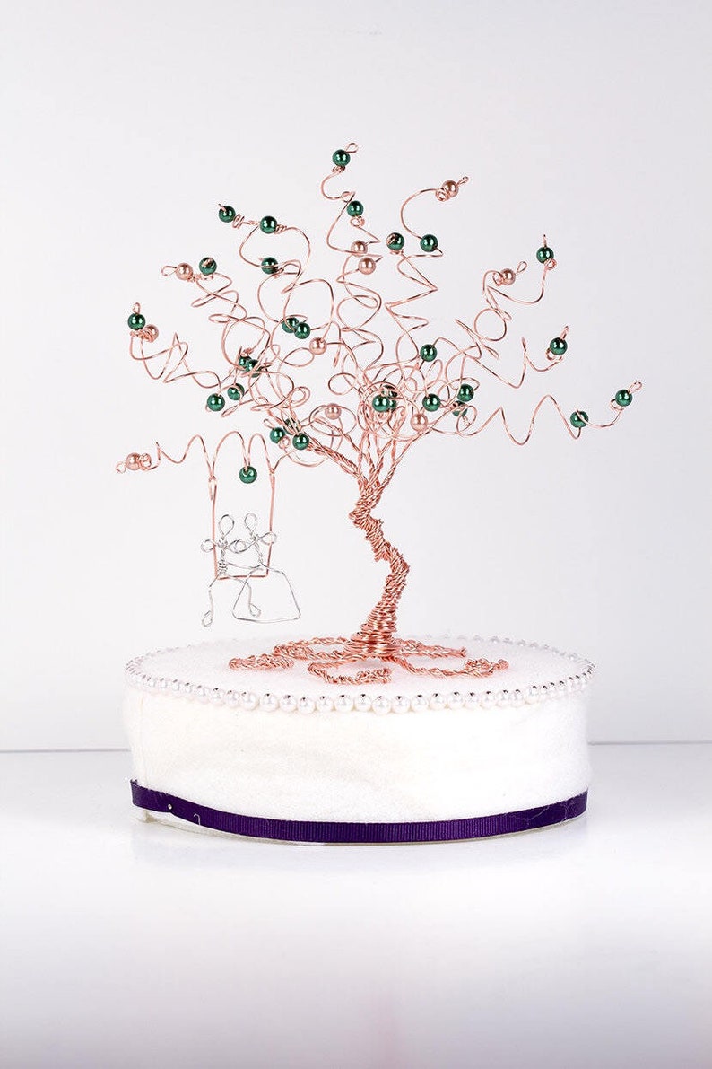 May include: A copper wire tree sculpture with green beads and a wire couple on a swing. The tree is on a white base with a purple ribbon.