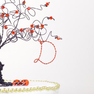Puede incluir: Una escultura de &aacute;rbol de alambre negro con cuentas de vidrio naranja y rojo. El &aacute;rbol est&aacute; decorado con un c&iacute;rculo de alambre de cobre y dos peque&ntilde;as calabazas naranjas con caras negras en la base.