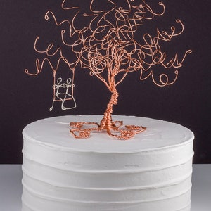 May include: A white cake with a copper wire tree topper. The tree has a small swing hanging from it.