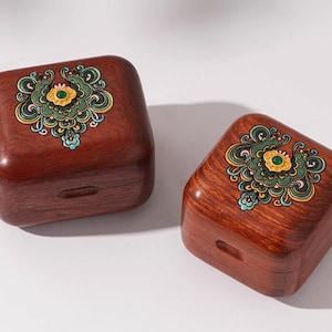 Puede incluir: Dos cajas cuadradas de madera en un rico color marrón rojizo. Cada caja presenta un diseño floral decorativo en tonos verdes, amarillos y azules. Las cajas tienen una pequeña abertura empotrada en la parte delantera. Probablemente para joyas o objetos pequeños.