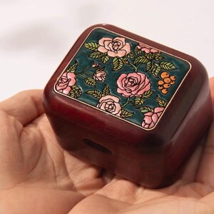 May include: A small, square wooden box with a dark red finish. The top features a teal panel with a floral design of pink roses and green leaves. The box is held in open hands.
