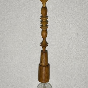 Op de afbeelding: Een hanglamp met een gedraaide houten steel met een reeks gestapelde, decoratieve geometrische vormen in een natuurlijke bruine afwerking. De lamp hangt aan een zwart snoer en eindigt in een heldere glazen bolvormige gloeilamp.