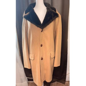 May include: A tan coat featuring a dark brown faux fur collar, long sleeves, and a front button closure with three visible buttons. The garment includes two front flap pockets positioned at the waist level, providing a structured appearance.