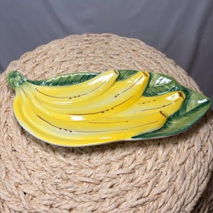 May include: A ceramic banana-shaped serving dish. The dish is painted yellow with brown accents to resemble bananas. The dish is set on a green leaf-shaped base. The dish is a decorative item for the kitchen or dining room.