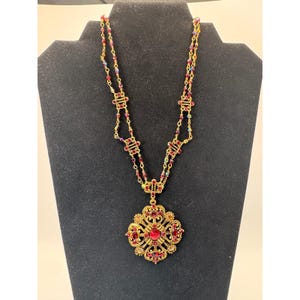 May include: A gold-tone necklace featuring a large, ornate, filigree-style pendant set with deep red and amber-colored crystals. The chain is composed of delicate gold links interspersed with small, multi-colored iridescent crystal beads.