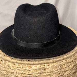 Puede incluir: Un sombrero fedora negro con una banda de cuero. El sombrero tiene una corona redondeada y un ala ancha. El sombrero está sobre una cesta tejida.