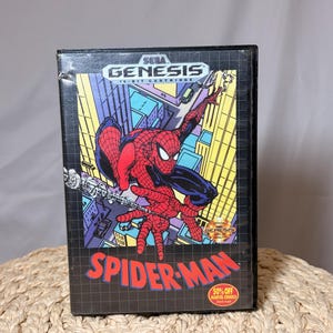 May include: A black Sega Genesis video game cartridge for Spider-Man. The box art features Spider-Man in a red and blue suit swinging through a city. The Sega Genesis logo is at the top, and the words "Spider-Man" are in red.