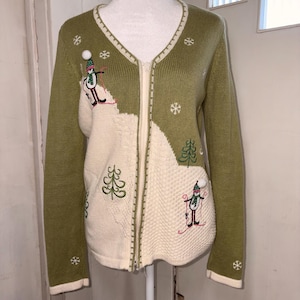 May include: Olive green zip-up sweater with a V-neck and embroidered winter scene. The sweater features a white textured design with embroidered snowmen, snowflakes, and a Christmas tree. The cuffs and neckline are trimmed in white.