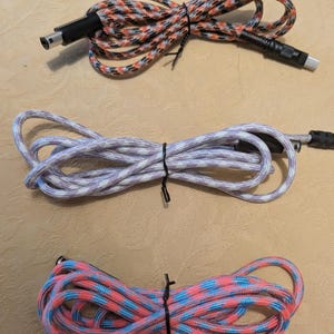May include: Three braided USB cables in the image. The top cable is orange, black, and white. The middle cable is white and purple. The bottom cable is pink and blue. All cables are tied with black ties.