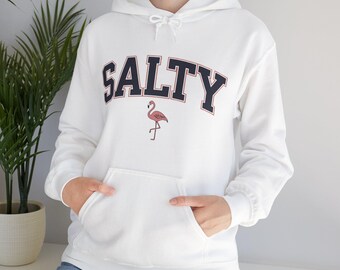 Könnte beinhalten: Weißer Kapuzenpullover mit Fronttasche und Kordelzugkapuze. Das Wort "SALTY" ist in marineblauen Blockbuchstaben mit einer rosa Flamingo-Grafik darunter auf die Brust gedruckt. Ein lässiges, bequemes Kleidungsstück.