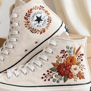 May include: Cream-colored high-top sneakers with floral embroidery. The sneakers feature white laces, a black stripe along the sole, and a circular patch with text and a star. The embroidered flowers include shades of orange, red, and green.