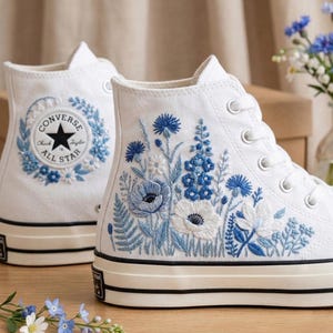 Puede incluir: Zapatillas altas blancas con diseños florales bordados en azul y blanco. Las zapatillas presentan el logotipo de Converse All Star en el lateral y una franja negra a lo largo de la suela. El bordado floral incluye varios tipos de flores y follaje.
