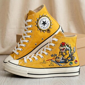 Puede incluir: Zapatillas altas Converse Chuck Taylor All Star de color amarillo mostaza. Un zapato presenta un personaje de Bowser conduciendo un kart, con banderas a cuadros y pieles de pl&aacute;tano. El otro zapato tiene un logotipo de Converse con el texto "All Star".